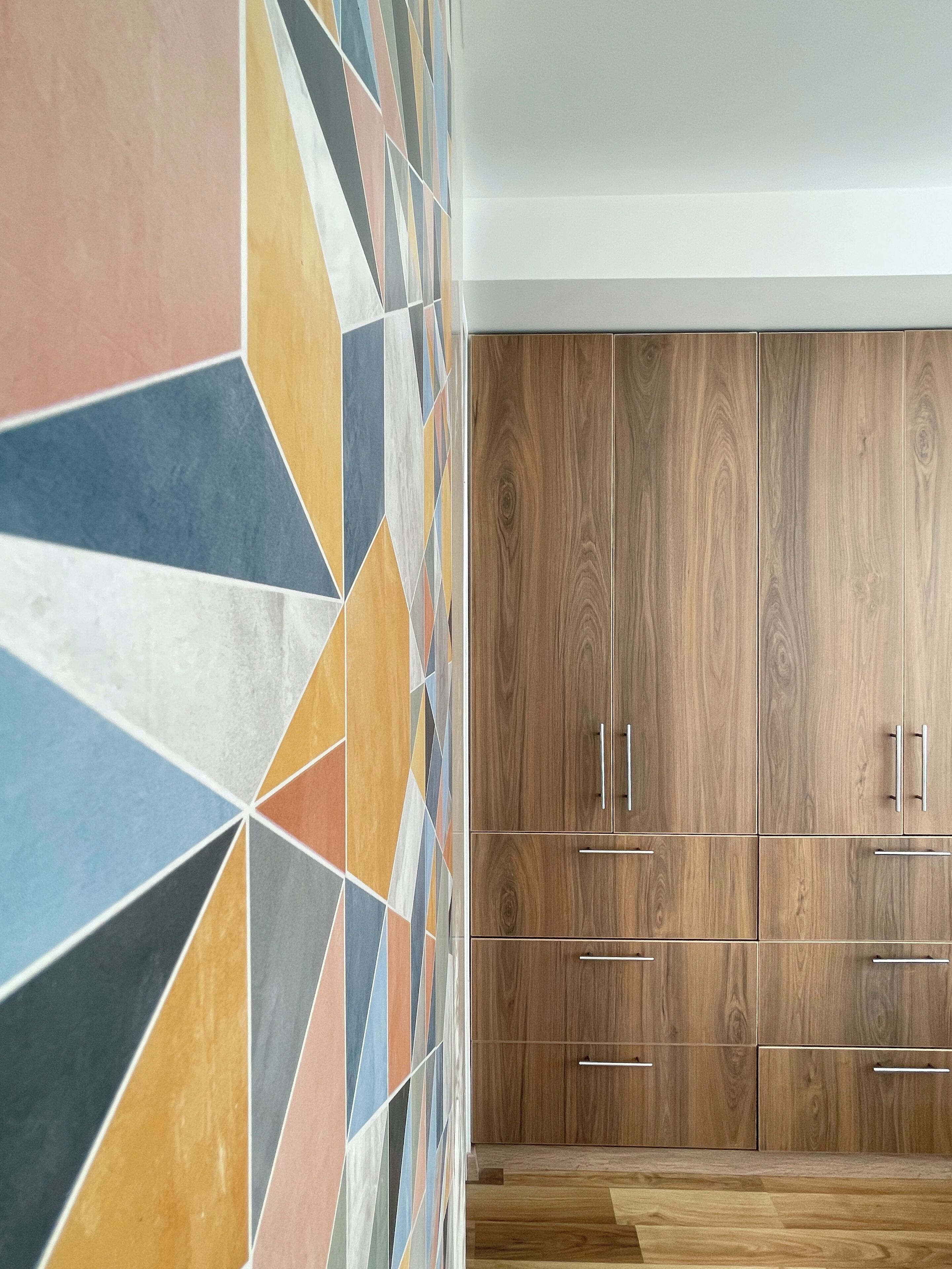Une pièce avec un mur orné d’un motif géométrique, des meubles en bois dotés de poignées métalliques et un plancher en bois.