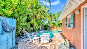 A backyard with a swimming pool, two Adirondack chairs, and a tiki-style umbrella. - Catching Waves – Coastal Retreat w/ Heated Pool, Steps to Beach, Pet Friendly (Holmes Beach)