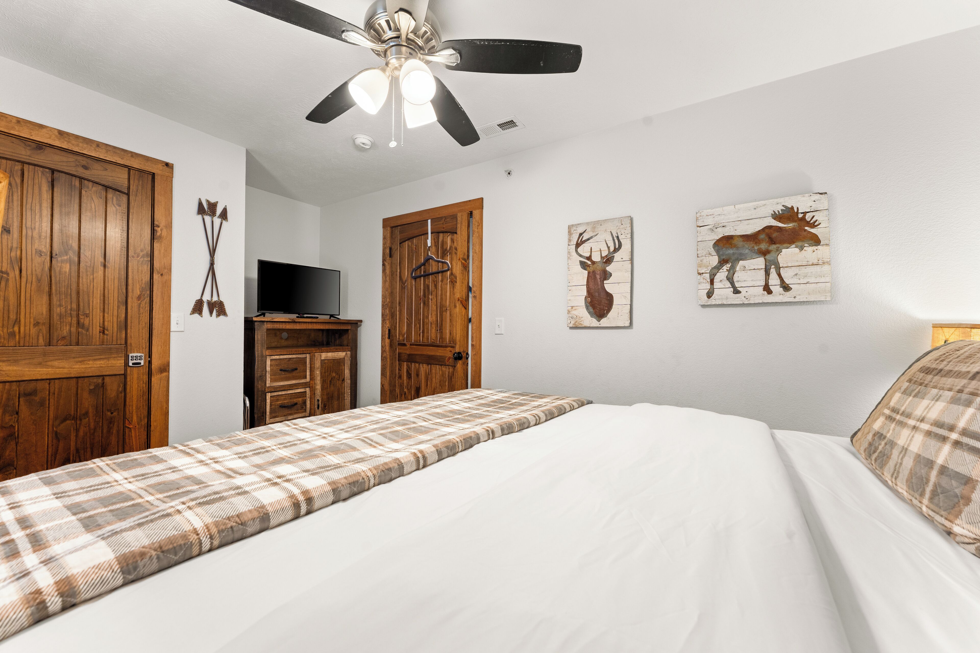 A bedroom with a bed, a ceiling fan, wooden doors, a television, and wall art.