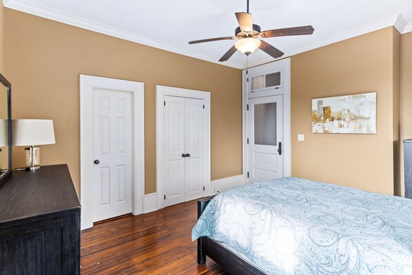 A bedroom with a bed, a ceiling fan, two white doors, a nightstand with a lamp, and a wall-mounted painting. - Train Depot | 2/1.5 | Heart of Downtown | Washer/Dryer (Natchez)