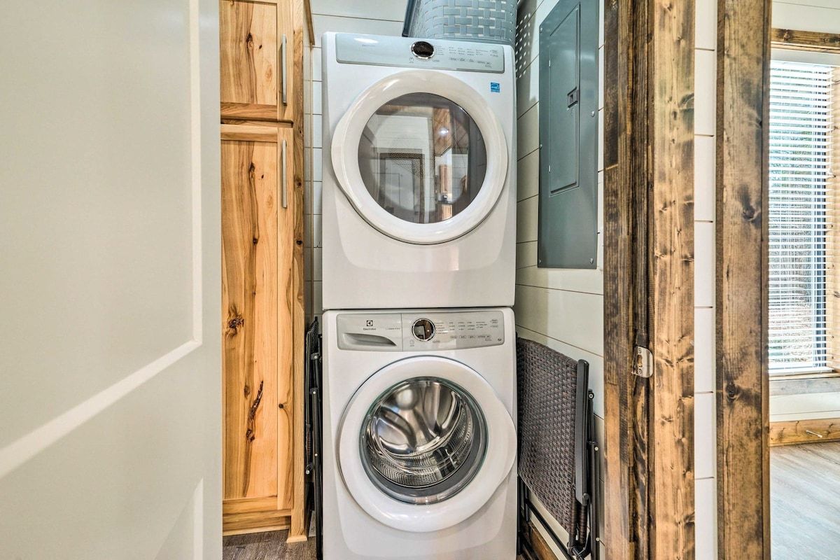 Une machine à laver et un sèche-linge superposés dans une buanderie, avec des armoires en bois et une fenêtre.