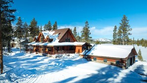 Ein großes Chalet mit schneebedecktem Dach, umgeben von hohen Kiefern und blauem Himmel.