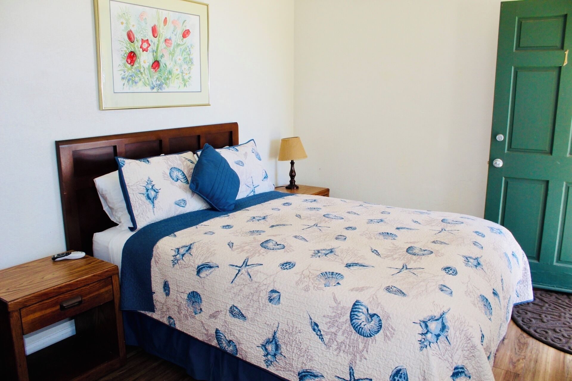 A neatly made bed with a floral print bedspread, a wooden nightstand with a lamp, and a green door.