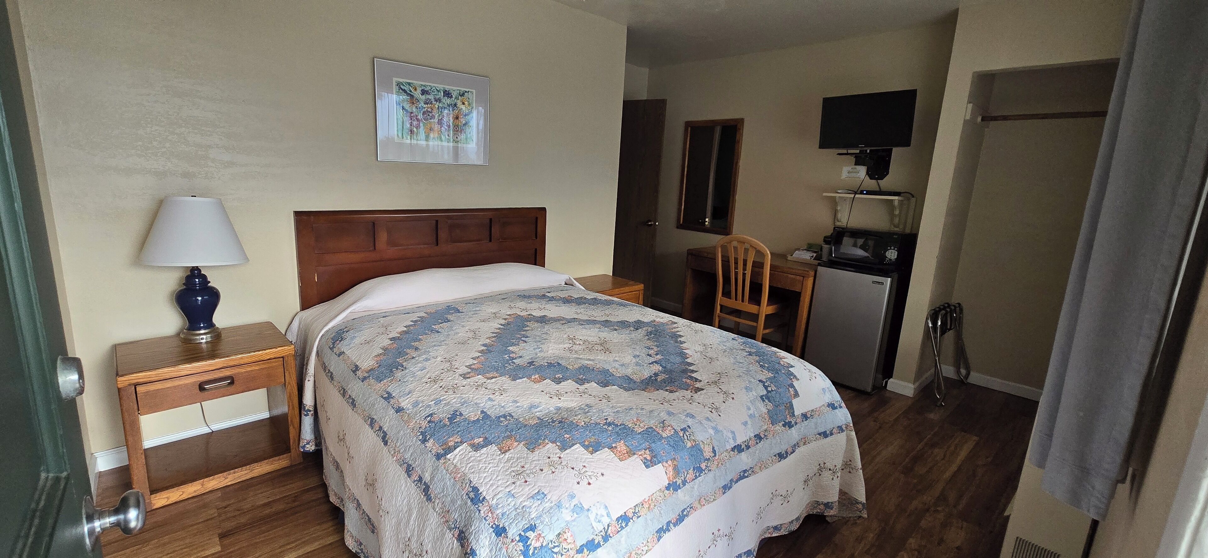 A hotel room with a bed, a nightstand with a lamp, a chair, a desk with a microwave, and a TV mounted on the wall.
