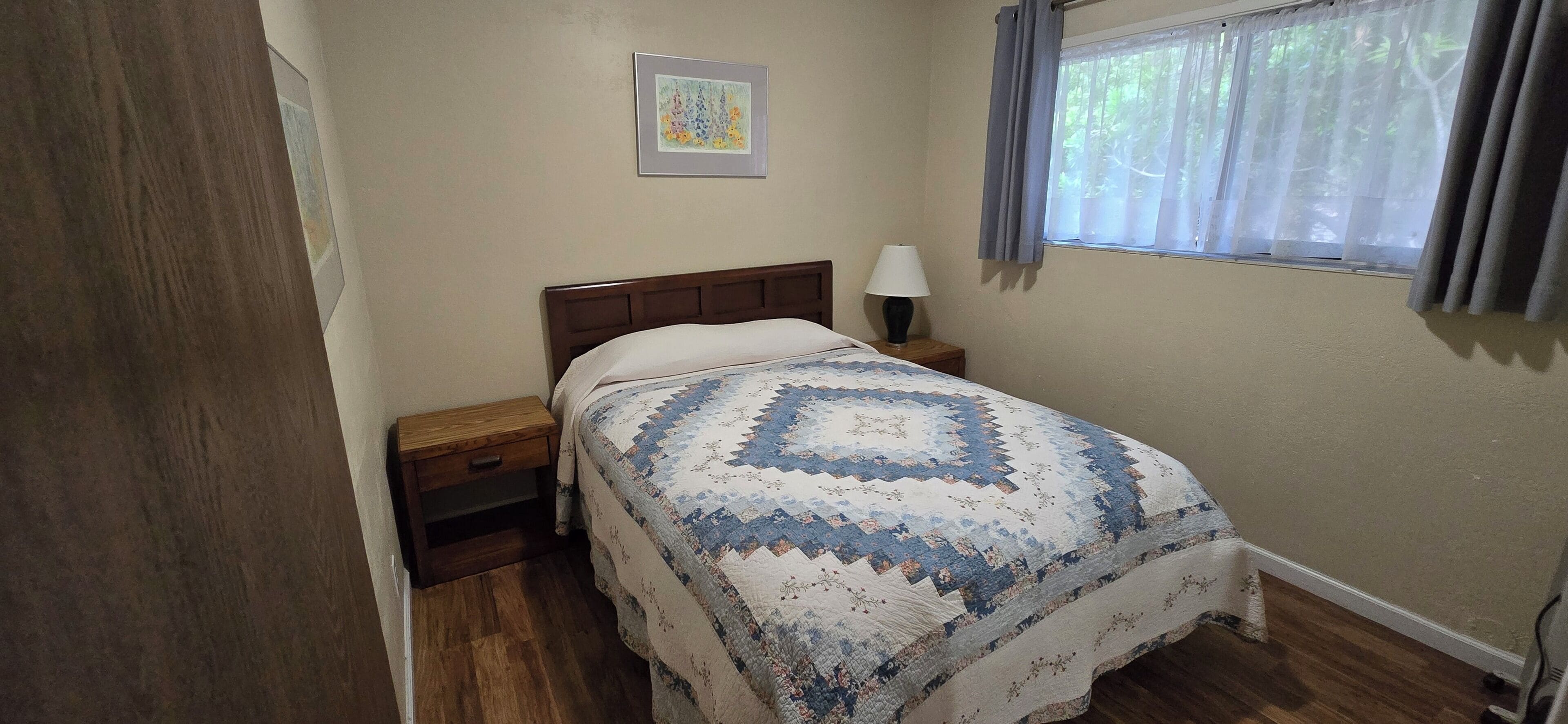 A bedroom with a bed, a nightstand, a lamp, a window with curtains, and a framed picture on the wall.