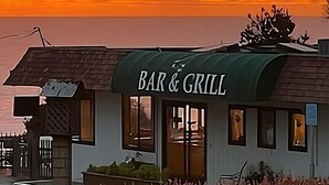 Un pequeño bar y restaurante con un letrero, cerca de un cuerpo de agua, y un atardecer en el fondo.