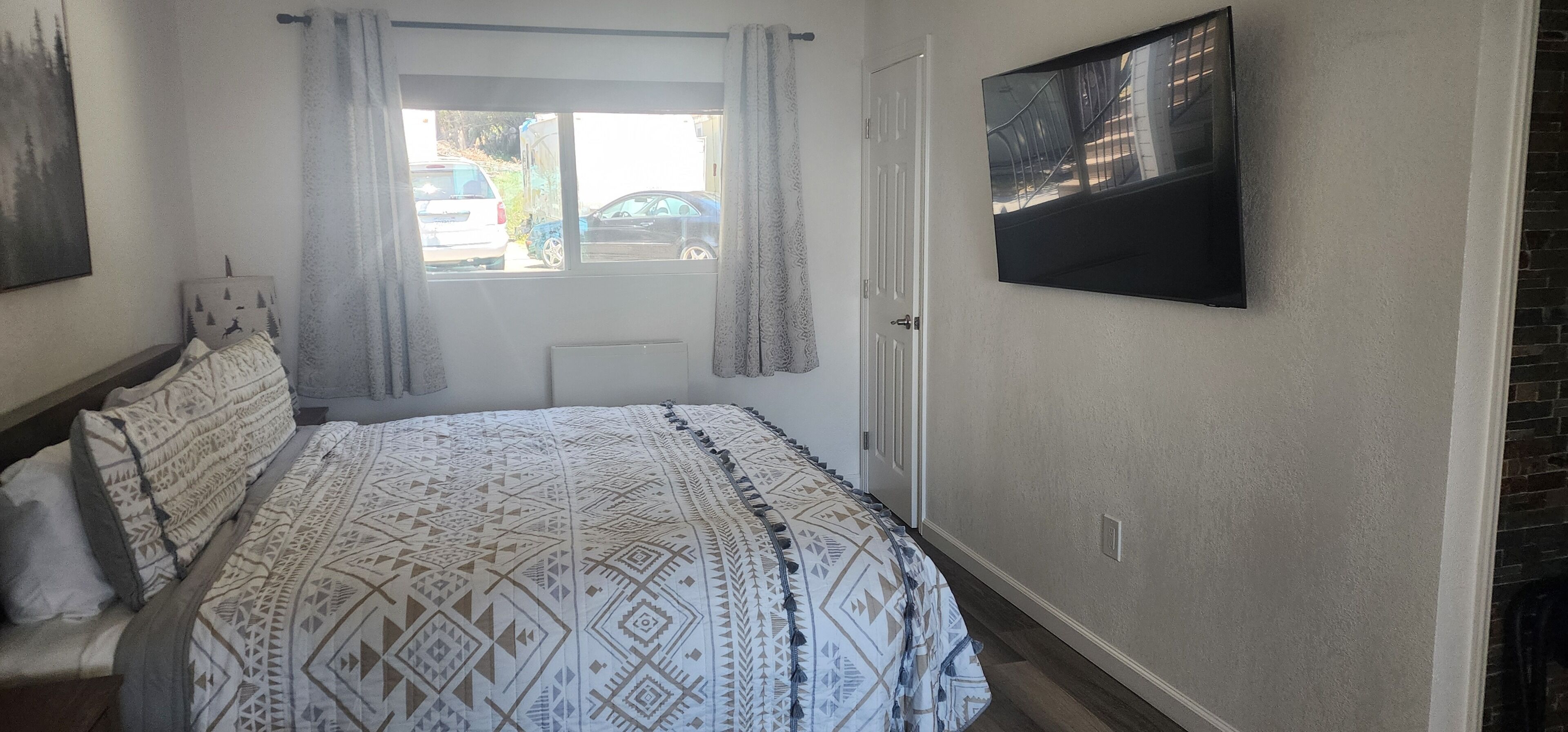 Un dormitorio con cama, ventana con cortinas, televisor montado y una puerta.
