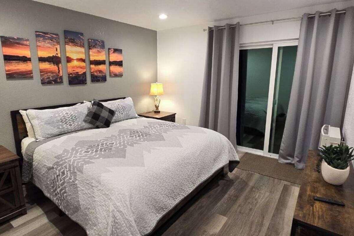 A modern bedroom with a large bed, two bedside lamps, a flat-screen TV, and a sliding glass door to a balcony.