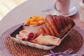 En frukostbricka med ägg, skinka, en croissant och frukt, placerat på en vävd bordsduk.