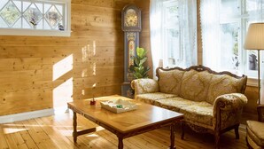 Ein Zimmer mit Holzwänden, einer großen, verzierten Uhr, einem gemusterten Sofa, einem Holztisch und einem Fenster mit Blick auf Grünflächen.