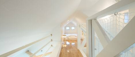 Un couloir étroit et bien éclairé, doté d’un plancher en bois et de murs blancs, qui mène à une chambre avec un lit et une fenêtre.