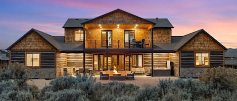 A two-story log cabin with a covered patio, a fire pit, and a view of the surrounding desert landscape.