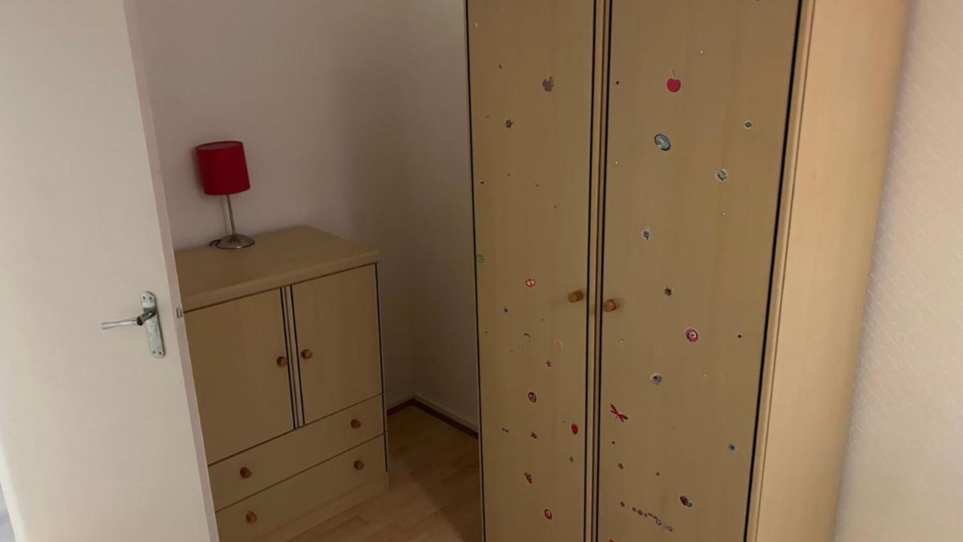 A room with a beige wardrobe, a small cabinet, and a red lamp on top.