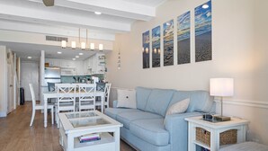 Una sala de estar moderna con un sofá azul claro, una mesa de centro de vidrio y una mesita de noche blanca con una lámpara. Se aprecia en el fondo una cocina con armarios blancos y una zona de comedor con mesa y sillas blancas.