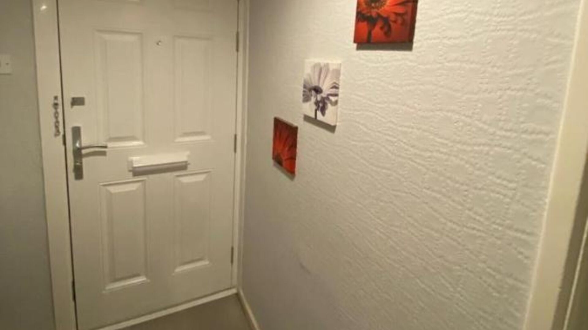 Un couloir avec une porte blanche, un mur blanc à texture et deux petits tableaux encadrés.