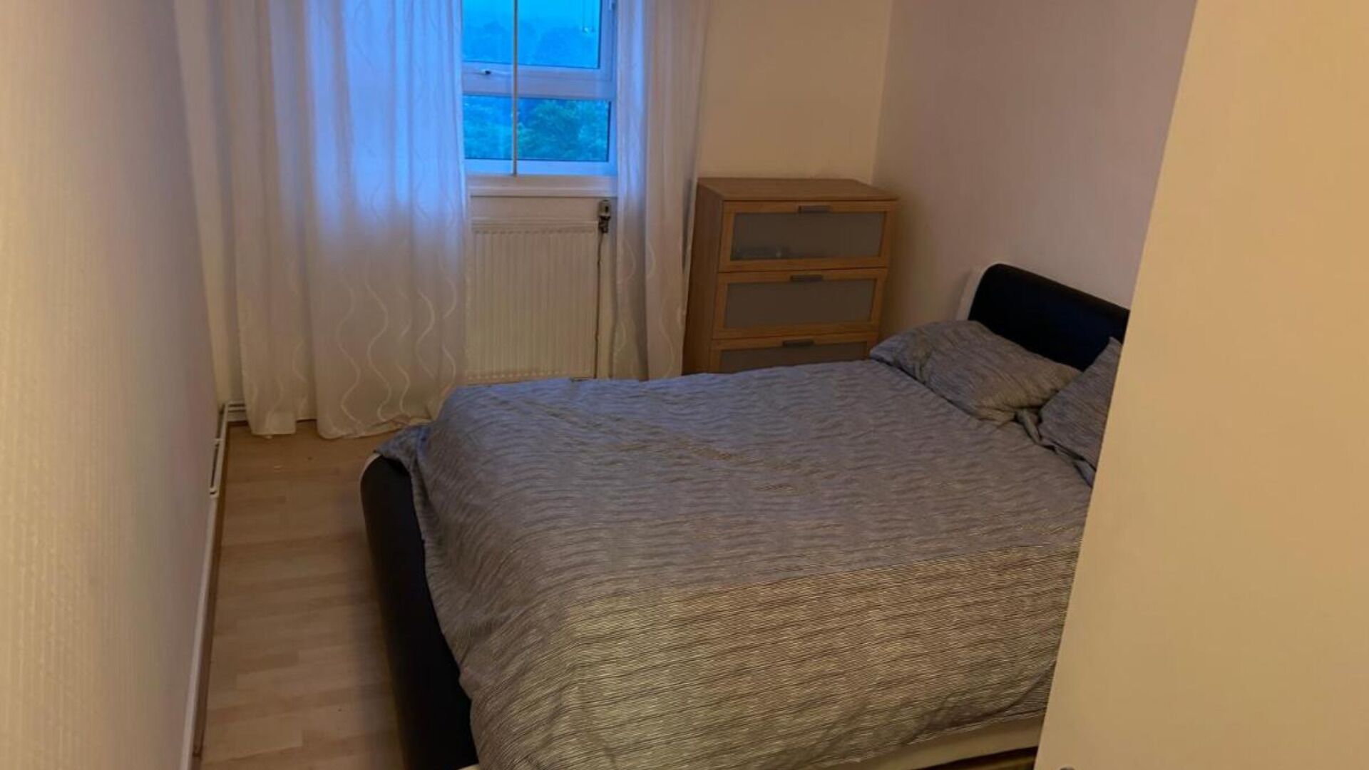 Ett litet sovrum med en säng, ett träbord som används som nattduksbord och ett fönster med gardiner.