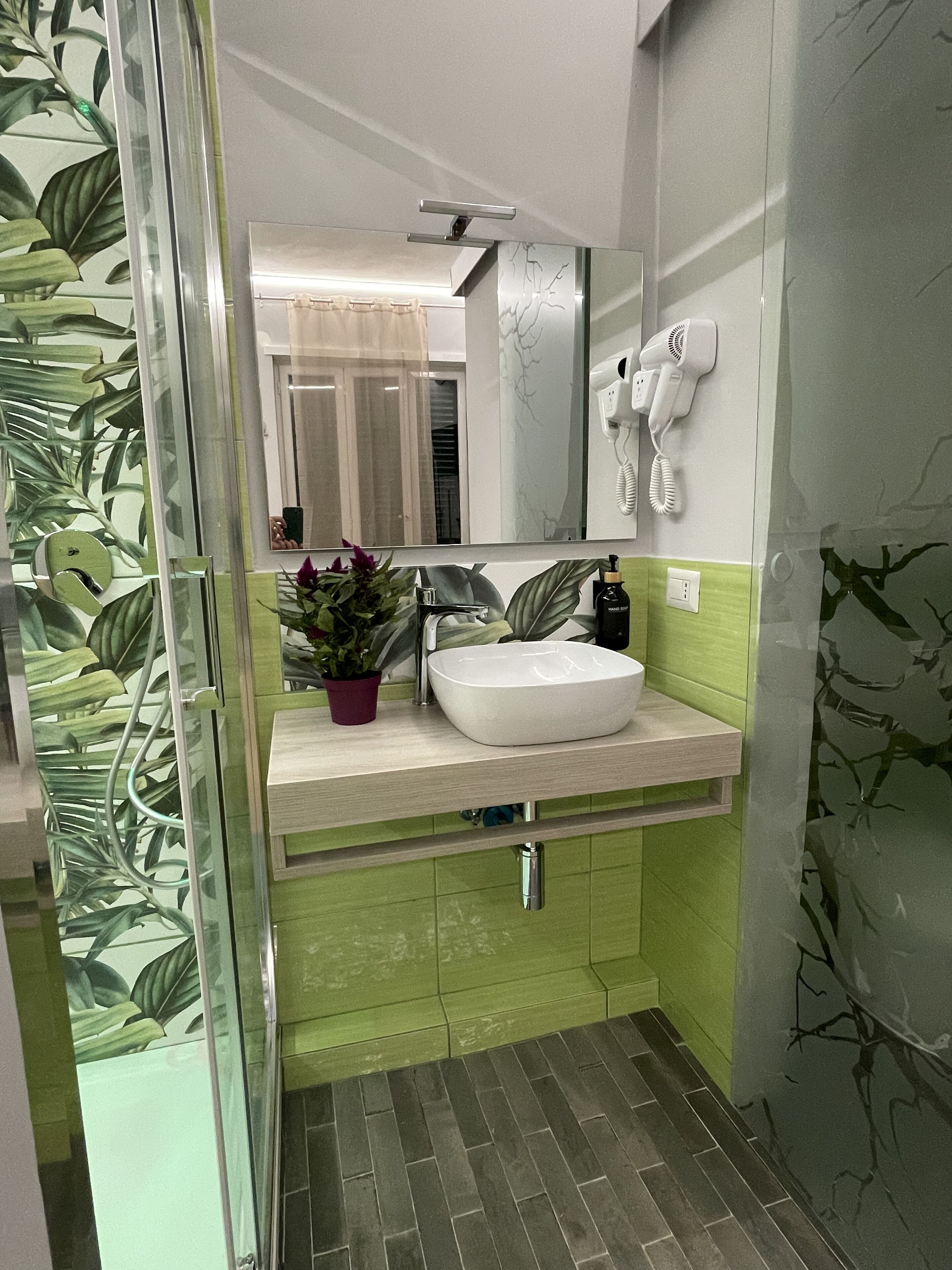 Ein Badezimmer mit einem weißen Waschbecken, grünen Wänden, einem Spiegel, einem Fön und einer Topfpflanze.