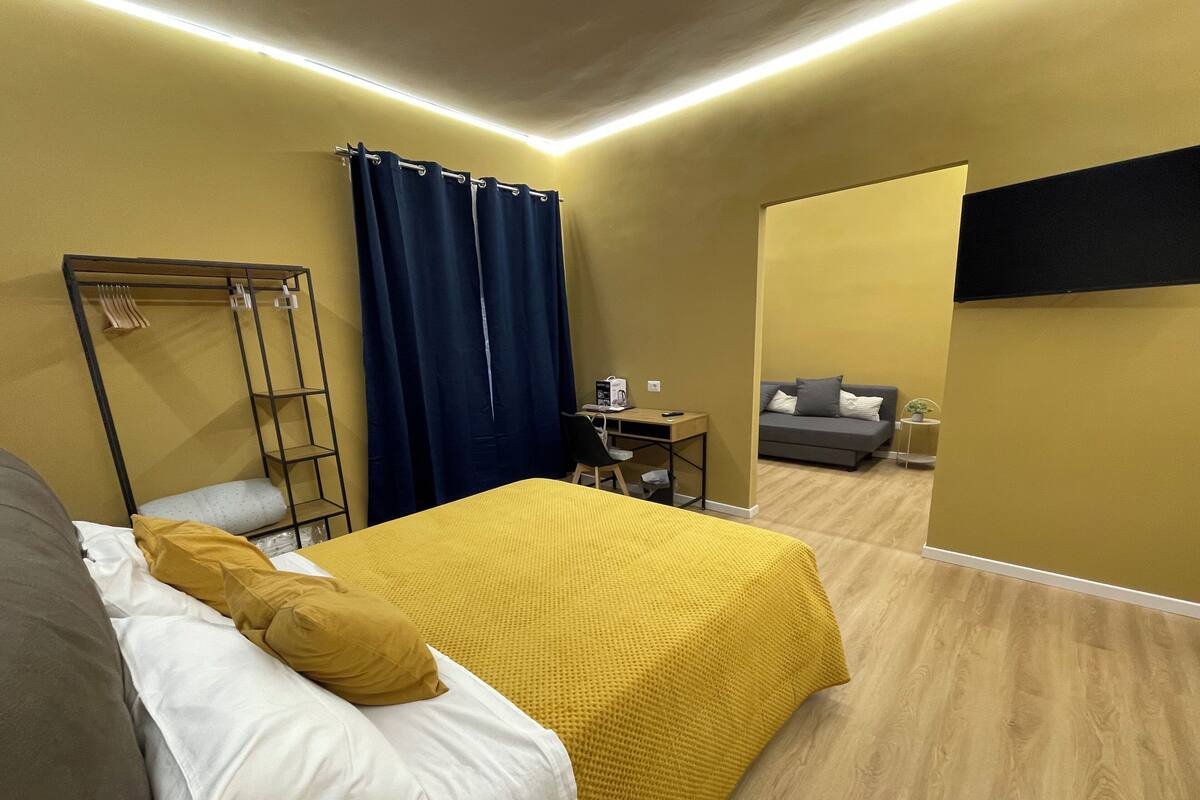 A modern hotel room with a bed, a desk, a sofa, and a TV mounted on the wall.