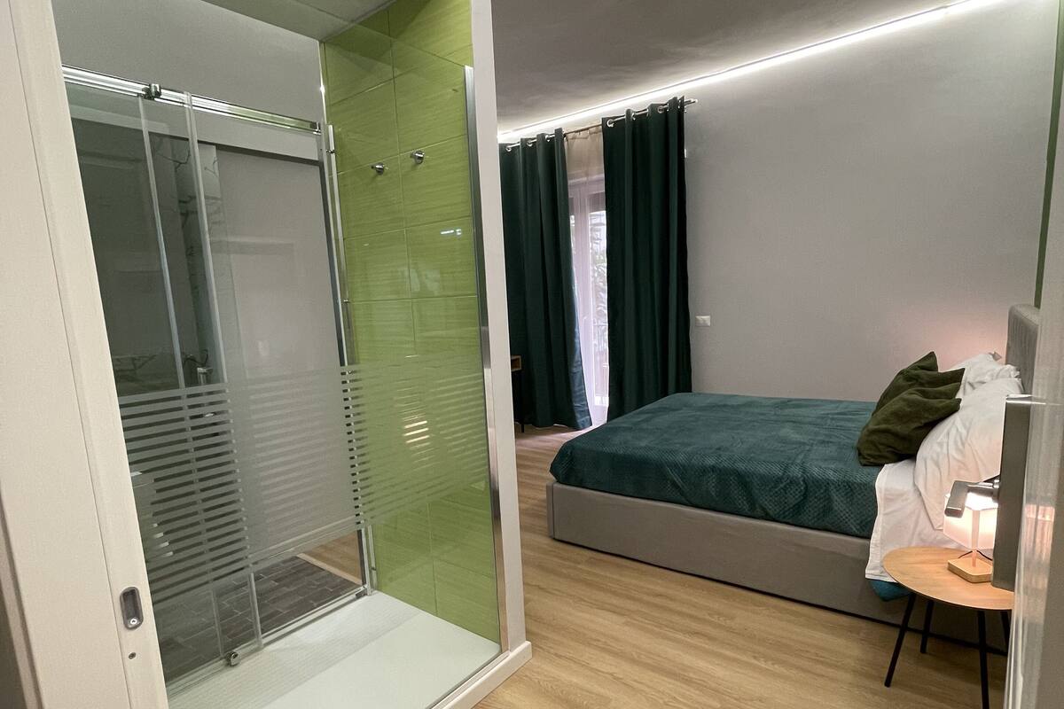 A modern bedroom with a glass shower enclosure, a bed with a green bedspread, and a bedside table with a lamp.