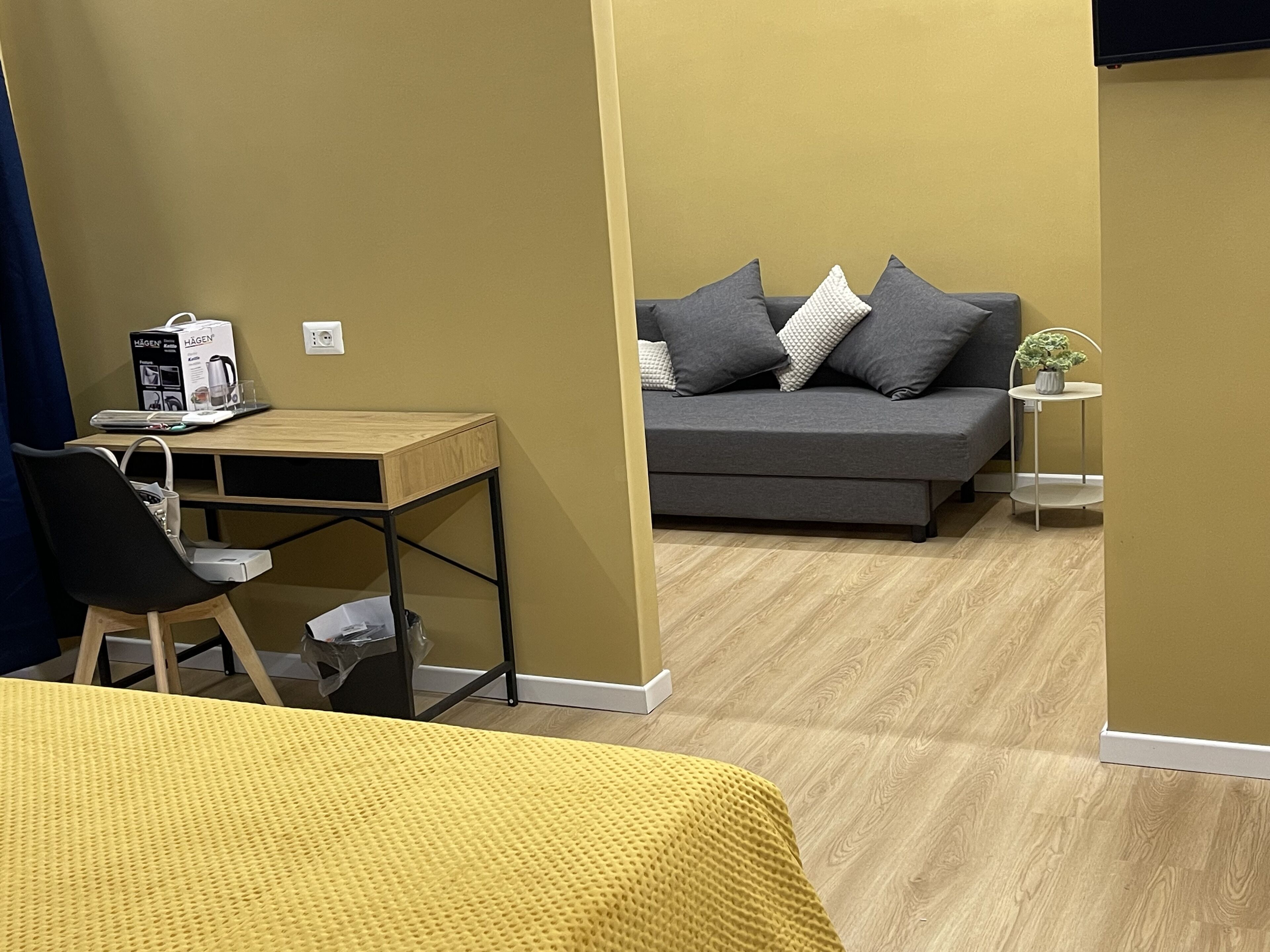 A room with a wooden desk, a grey sofa with pillows, a small white table with a plant, and a yellow bedspread.