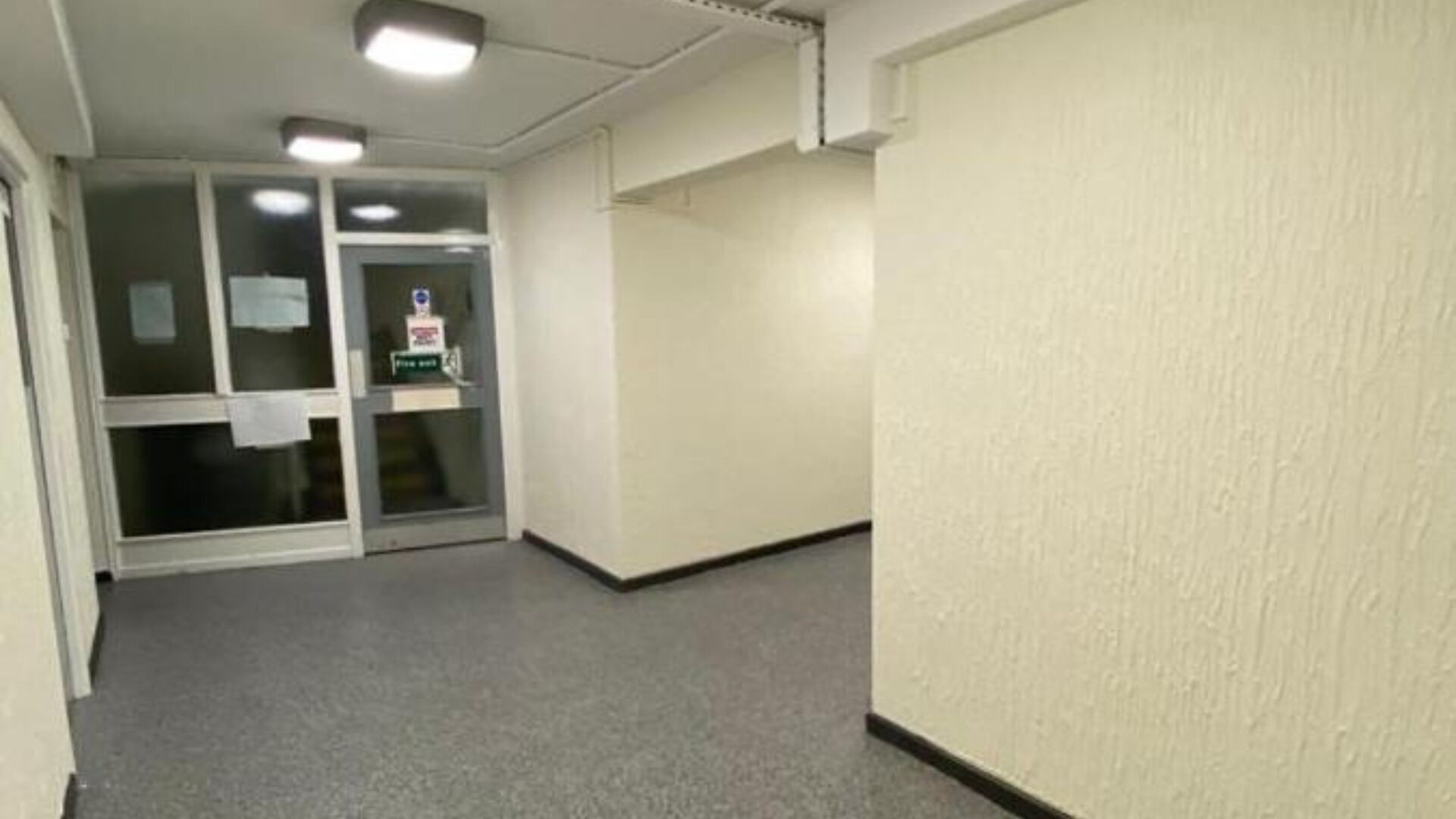 Un couloir avec une porte d’entrée en verre, un détecteur d’incendie et un luminaire au plafond.