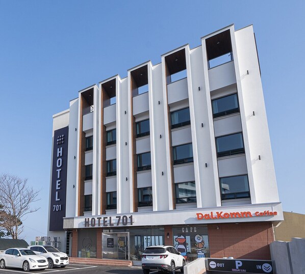 Un moderno edificio de hotel con un llamativo letrero que dice 'HOTEL 701', una cafetería llamada 'Dal.Komm Coffee' en la planta baja y varios coches estacionados al frente.