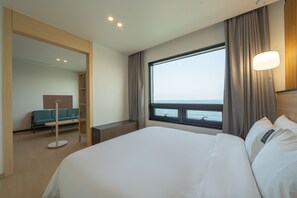 Habitación de hotel con una cama grande, zona de estar con sofá y mesa, y ventana con vista al mar.