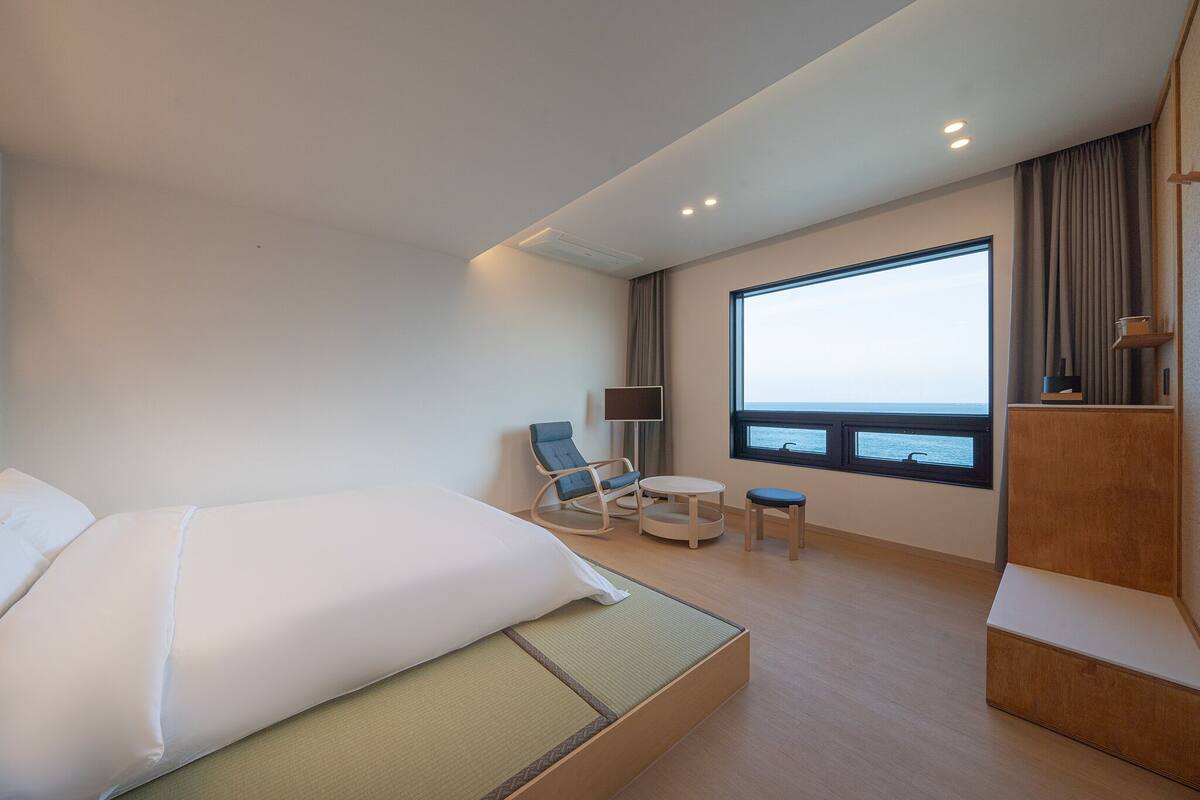 Una habitación de hotel moderna con una cama grande, una zona de estar con una silla y una mesa, un televisor y un ventanal con vistas.