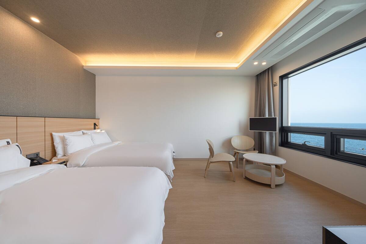 Habitación de hotel con dos camas, un escritorio, una silla, un televisor y un ventanal con vista al mar.