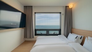 Une chambre d’hôtel dotée d’une grande fenêtre donnant sur la mer, d’un lit avec du linge de lit blanc et d’un téléviseur à écran plat fixé au mur.