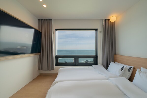 A hotel room with a large window offering a view of the sea, a bed with white linens, and a flat-screen TV mounted on the wall.