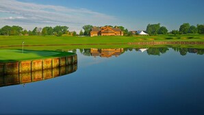 A golf course with a clubhouse, a pond reflecting the scene, and a clear sky.