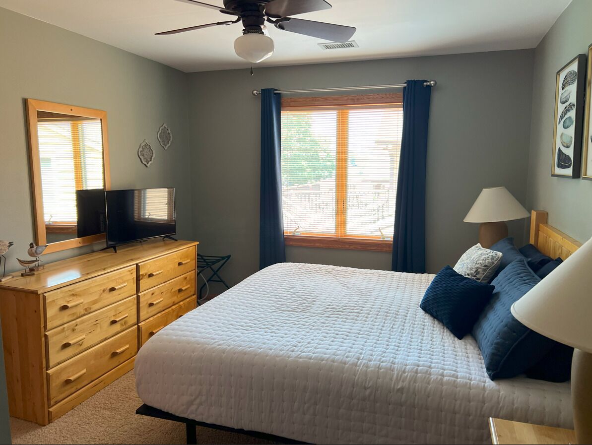 Une chambre à coucher avec un lit, une commode, un téléviseur et une fenêtre avec des rideaux.