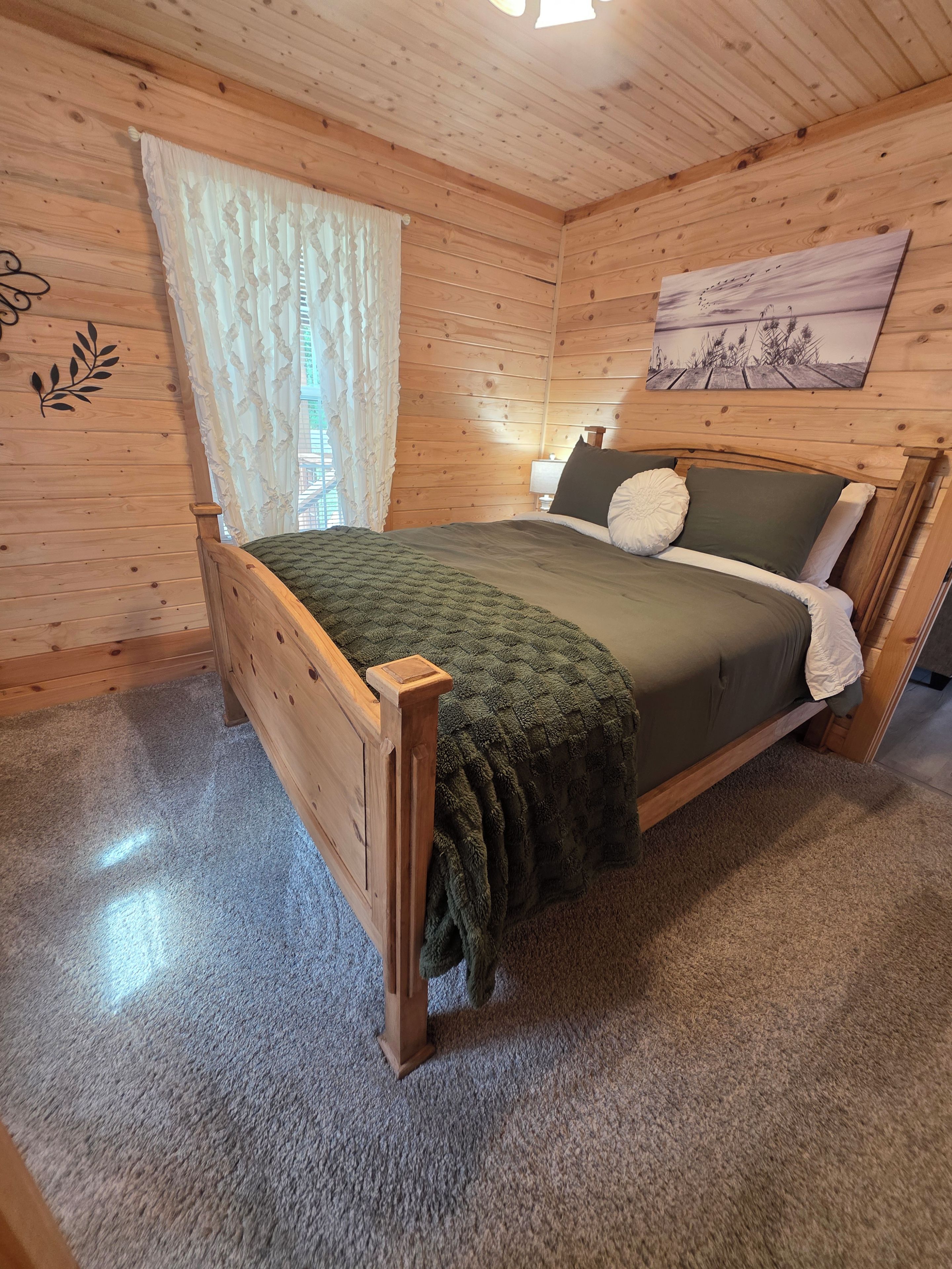 A wooden-paneled bedroom with a bed, a window with curtains, a wall-mounted painting, and a small bedside table.