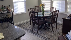Un coin repas avec une table ronde, des chaises et un lustre. On y trouve des fenêtres équipées de stores, une horloge au mur et un tapis sur le sol.