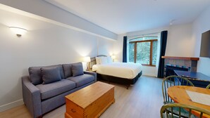 Moderni hotellihuone, jossa on harmaa sohva, puinen lipasto, valkoisilla lakanoiden päällä varustettu sänky, pieni pöytä tuoleineen ja suuri ikkuna, jossa on verhot.