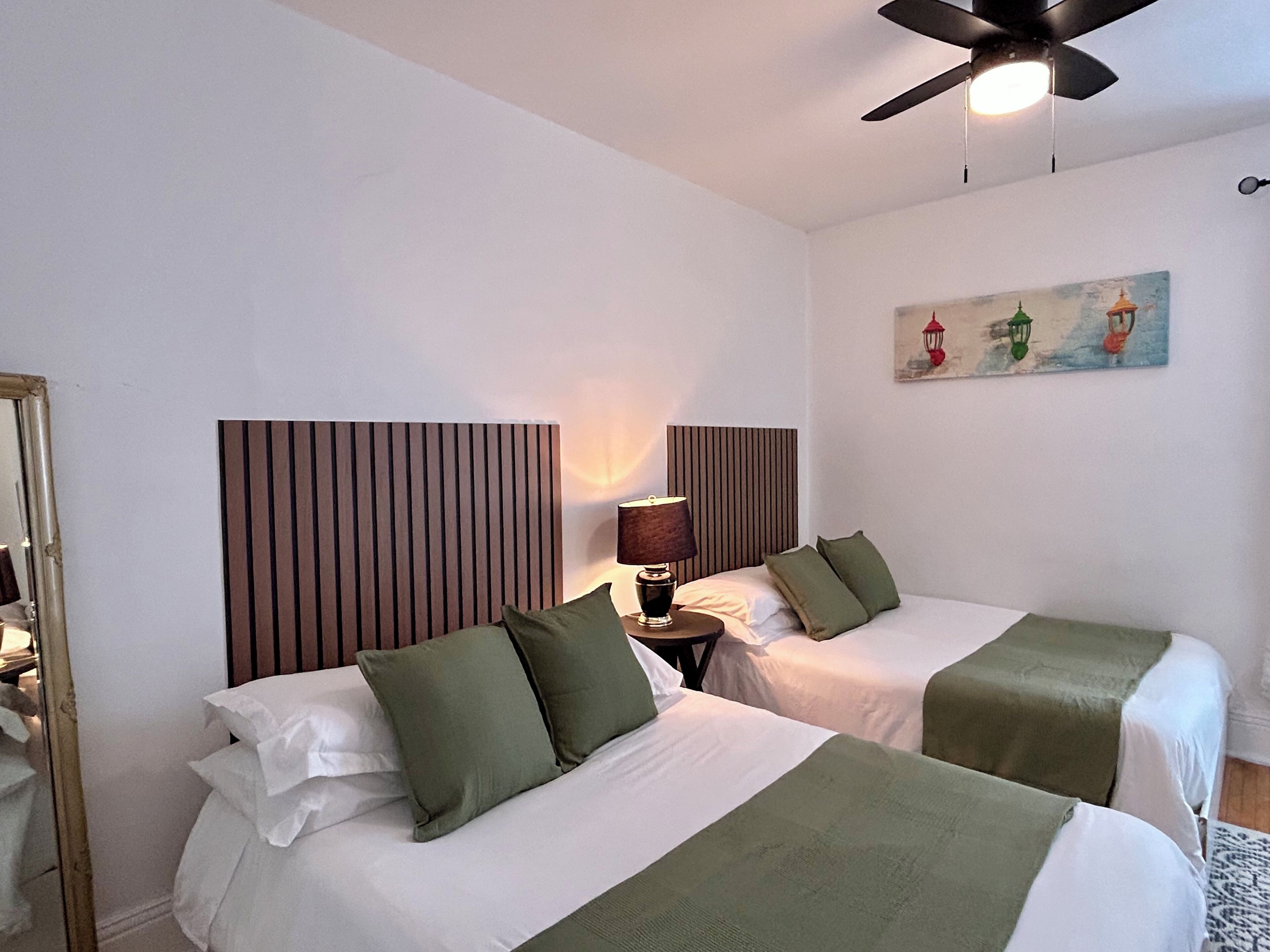 Une chambre d’hôtel avec deux lits, un ventilateur de plafond, une table de chevet avec une lampe et un tableau au mur.