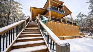 Un escalier en bois mène à un chalet à plusieurs niveaux, doté de balcons et entouré d’arbres couverts de neige.