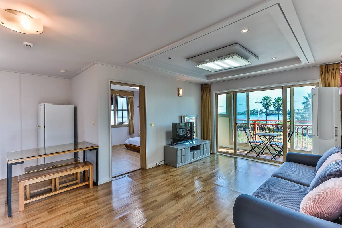 Un vaste espace de vie doté d’un plancher en bois, d’un canapé gris, d’un meuble pour la télévision et d’un balcon aménagé avec du mobilier d’extérieur.