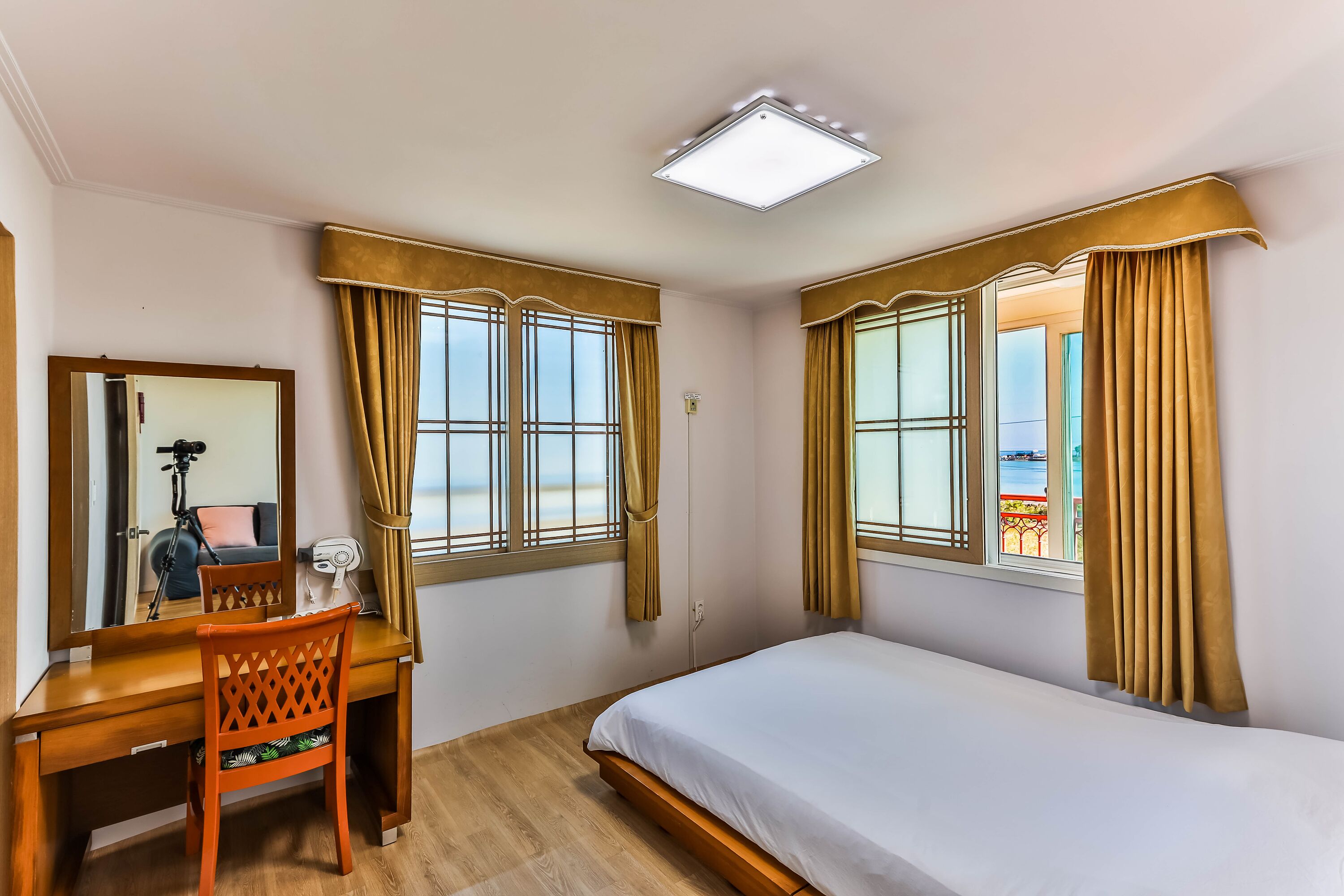 Une chambre à coucher avec un lit, un bureau et une chaise. Il y a des fenêtres avec des rideaux, un luminaire au plafond et une vue sur l’extérieur.