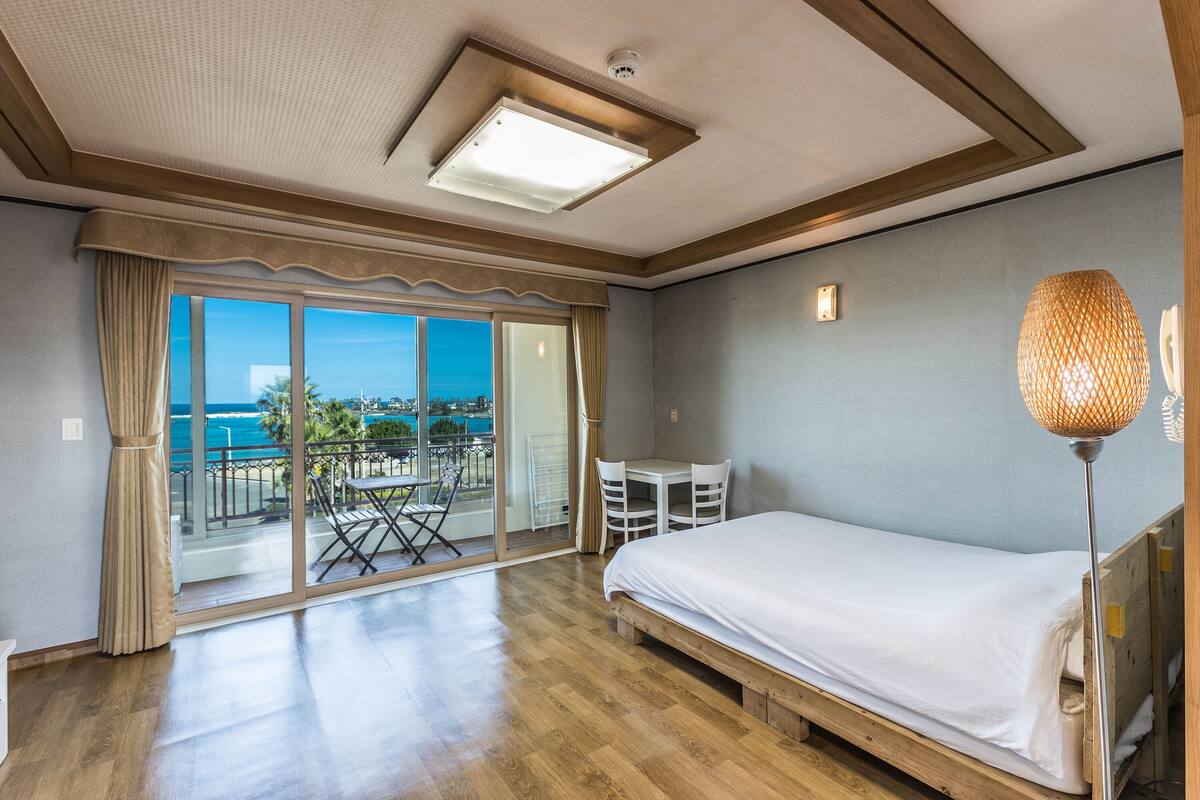Une chambre d’hôtel avec un lit, une table, des chaises, un balcon donnant sur la mer et un luminaire au plafond.