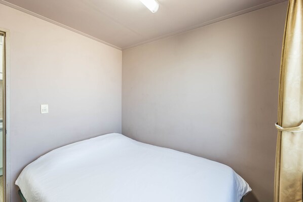 A small, single-bed room with a white bedspread, beige walls, and a ceiling light. - Sea Eco Beach (Jeju City)