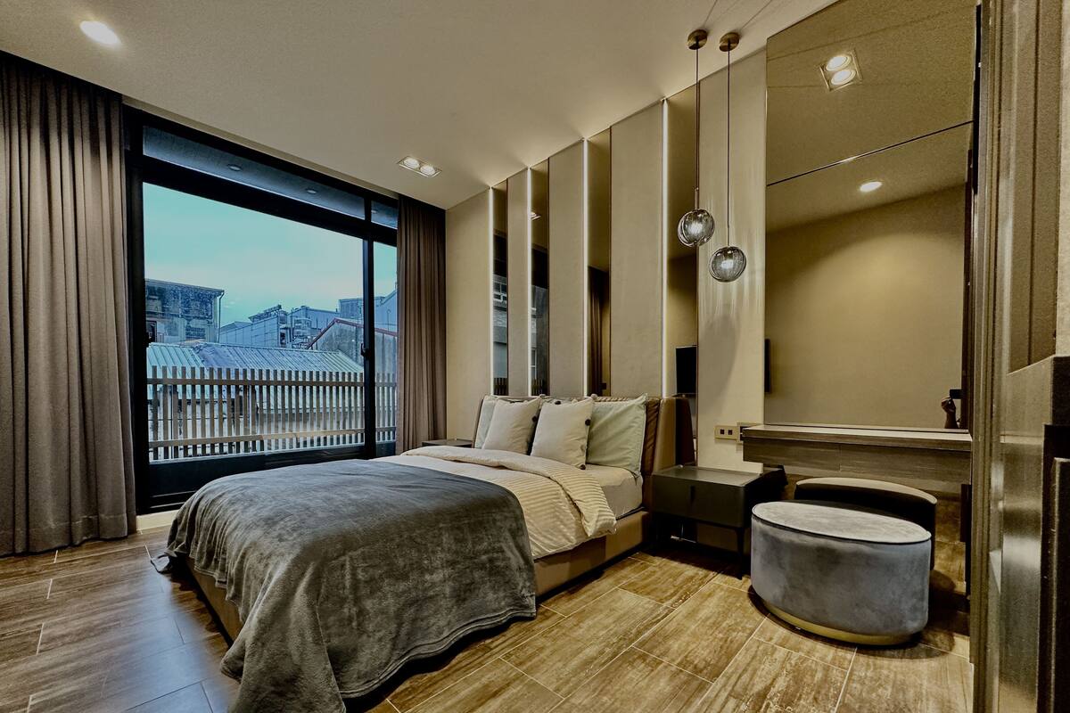Ein modernes Schlafzimmer mit einem großen Bett, einem Nachttisch, einem Spiegel und Blick auf die Stadt durch ein großes Fenster.