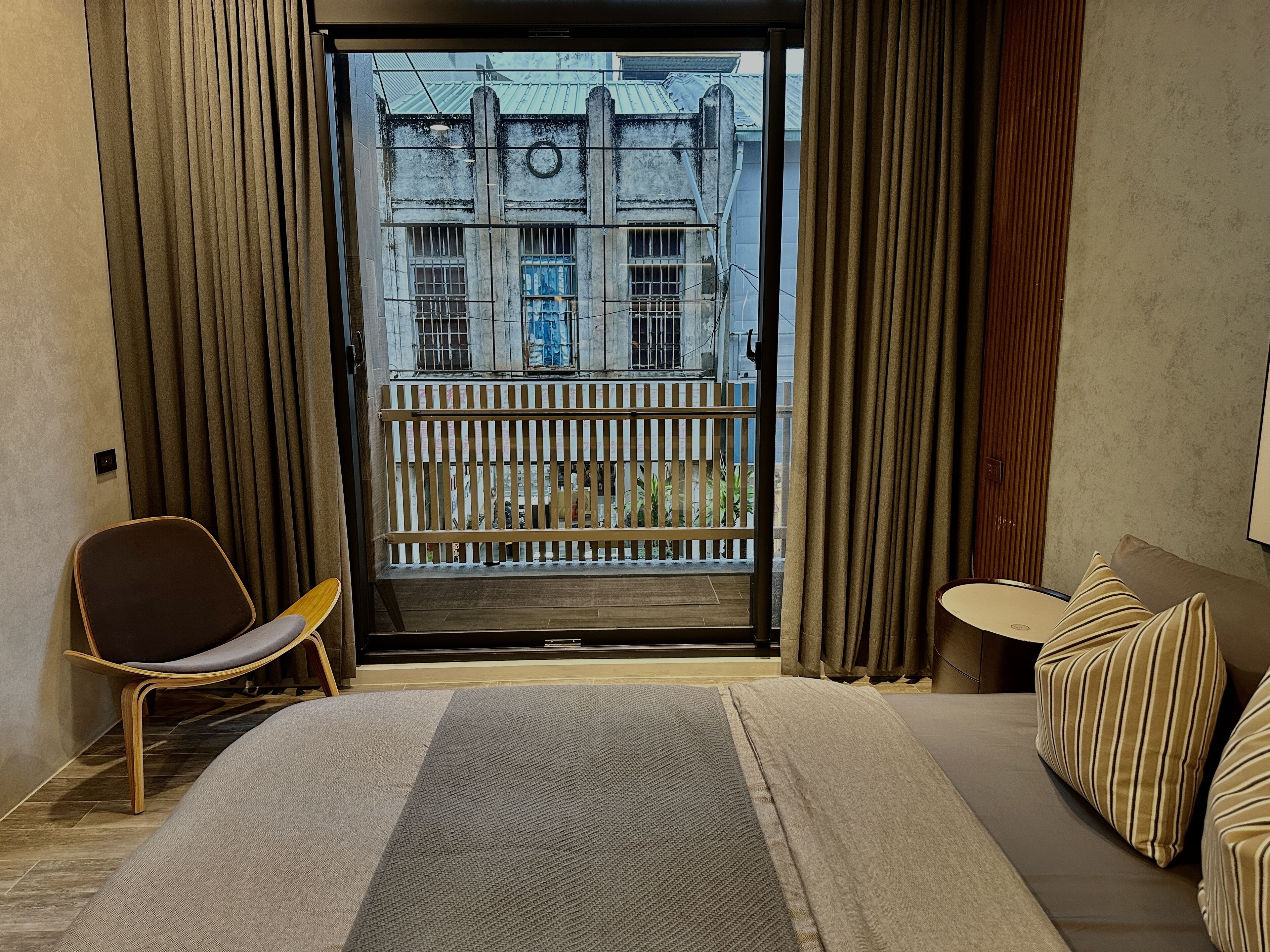 Une chambre d’hôtel avec un lit, une chaise, une petite table et une vue sur un immeuble, visible par la porte d’un balcon.