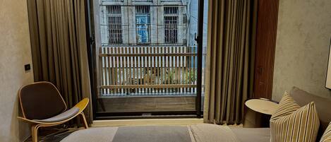 Une chambre d’hôtel avec un lit, une chaise, une petite table et une vue sur un immeuble, visible par la porte d’un balcon.