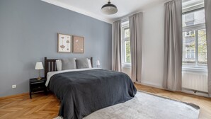 A bedroom with a bed, two bedside tables, a lamp, a large window with curtains, and artwork on the wall.