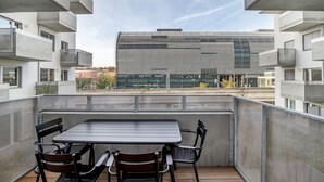 Ein Balkon mit Tisch und Stühlen, mit Blick auf ein modernes Gebäude mit horizontalen Linien und großen Fenstern.