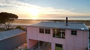 Vaaleanpunainen rakennus, jonka katto on tehty metallista, sijaitsee lähellä rantaa, ja taustalla näkyy auringonlasku.