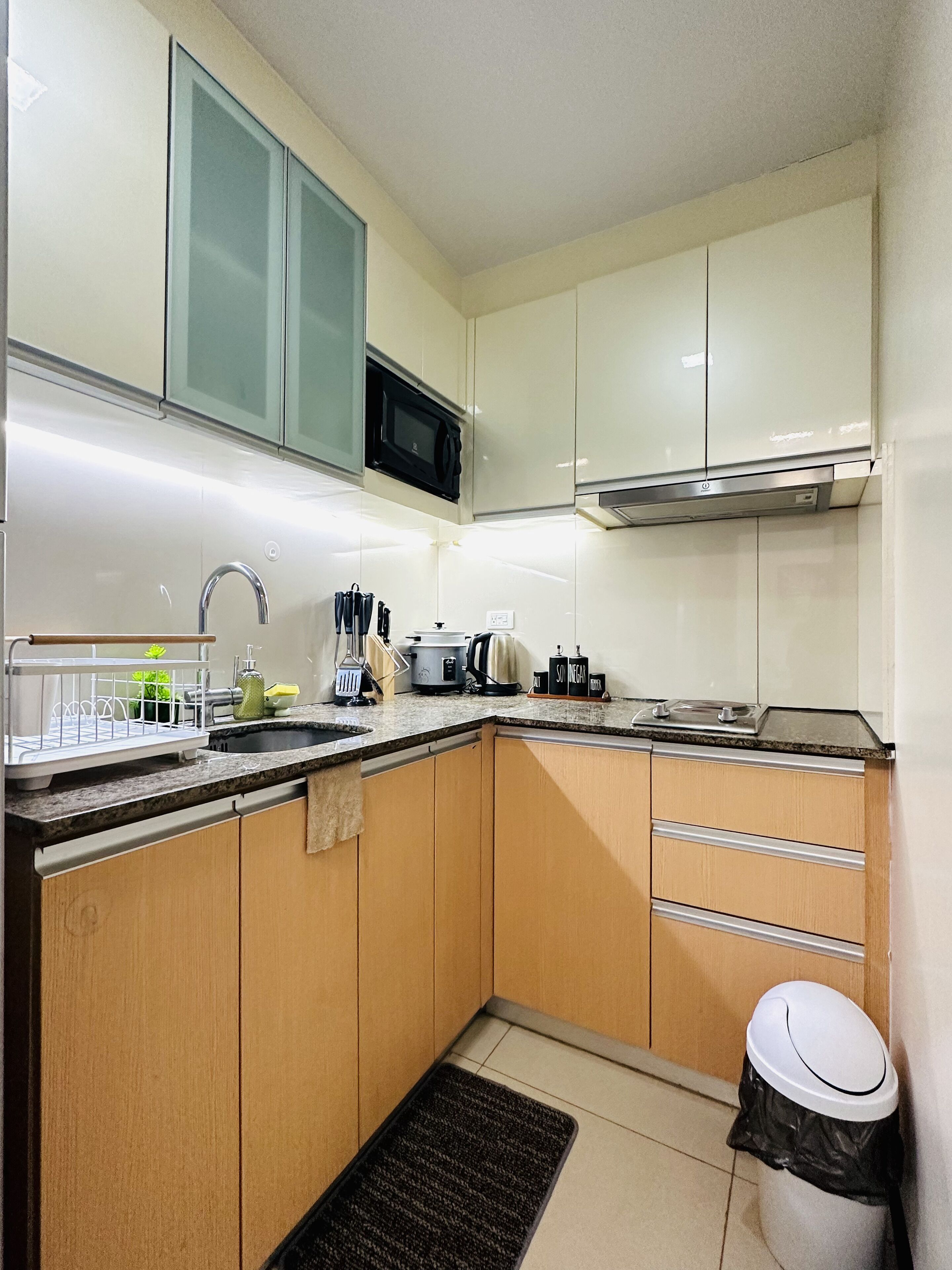 A compact kitchen with a microwave, stove, and sink. There are wooden cabinets and a black trash bin.
