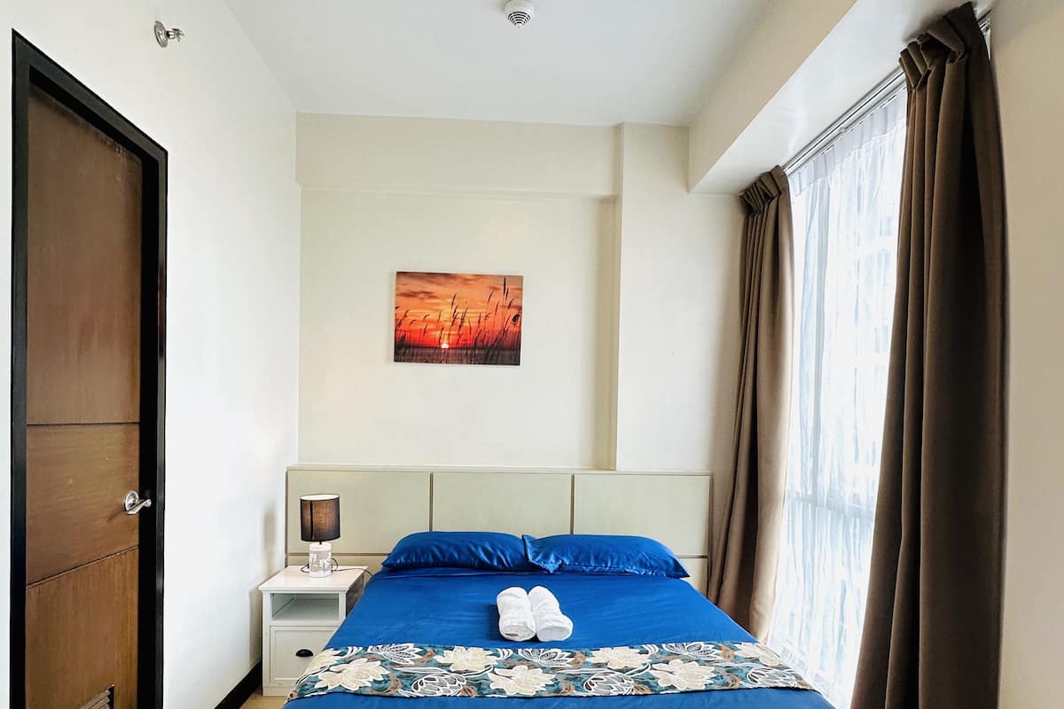 Una cama bien hecha con ropa de cama azul, una mesita de noche con lámpara, un cuadro en la pared y una puerta a la izquierda.